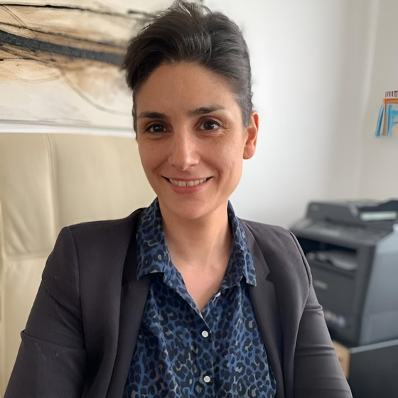 Une personne souriante aux cheveux courts et foncés, portant une chemise bleue à motifs et un blazer anthracite, dans un environnement de bureau.