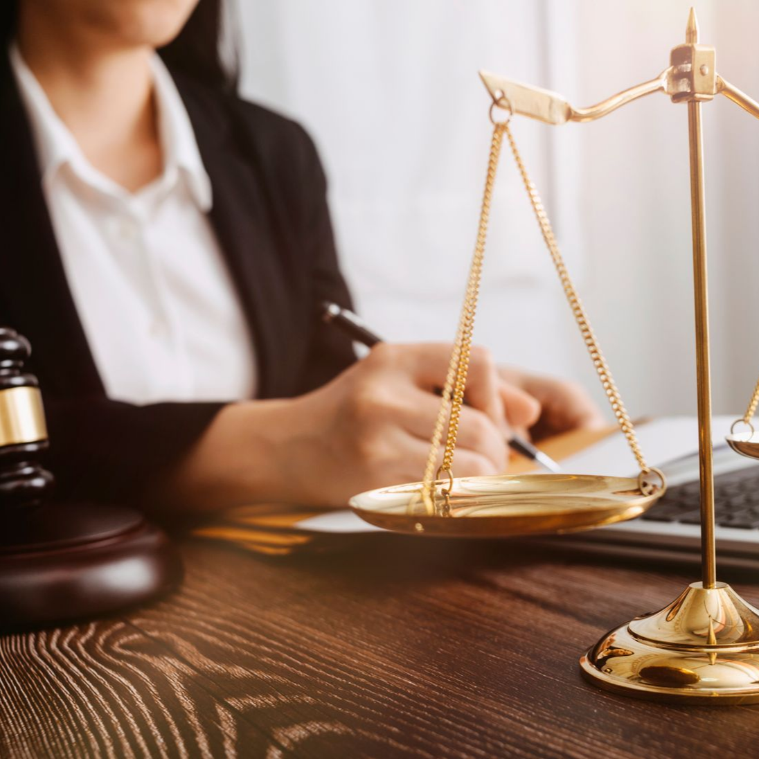 Une personne en costume écrit tandis qu'une balance de la justice et un maillet sont posés sur un bureau en bois.