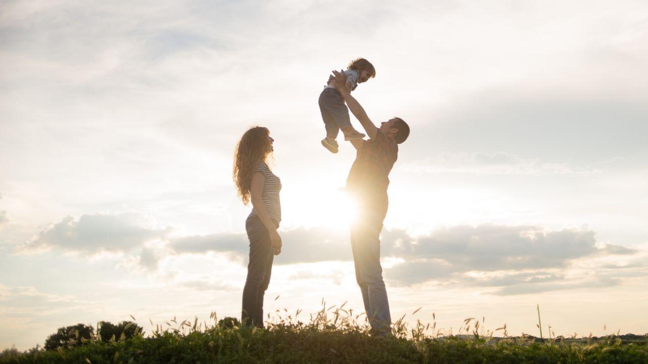 Une famille se découpe en silhouette sur un ciel lumineux : le père lance l'enfant en l'air, la mère regarde.