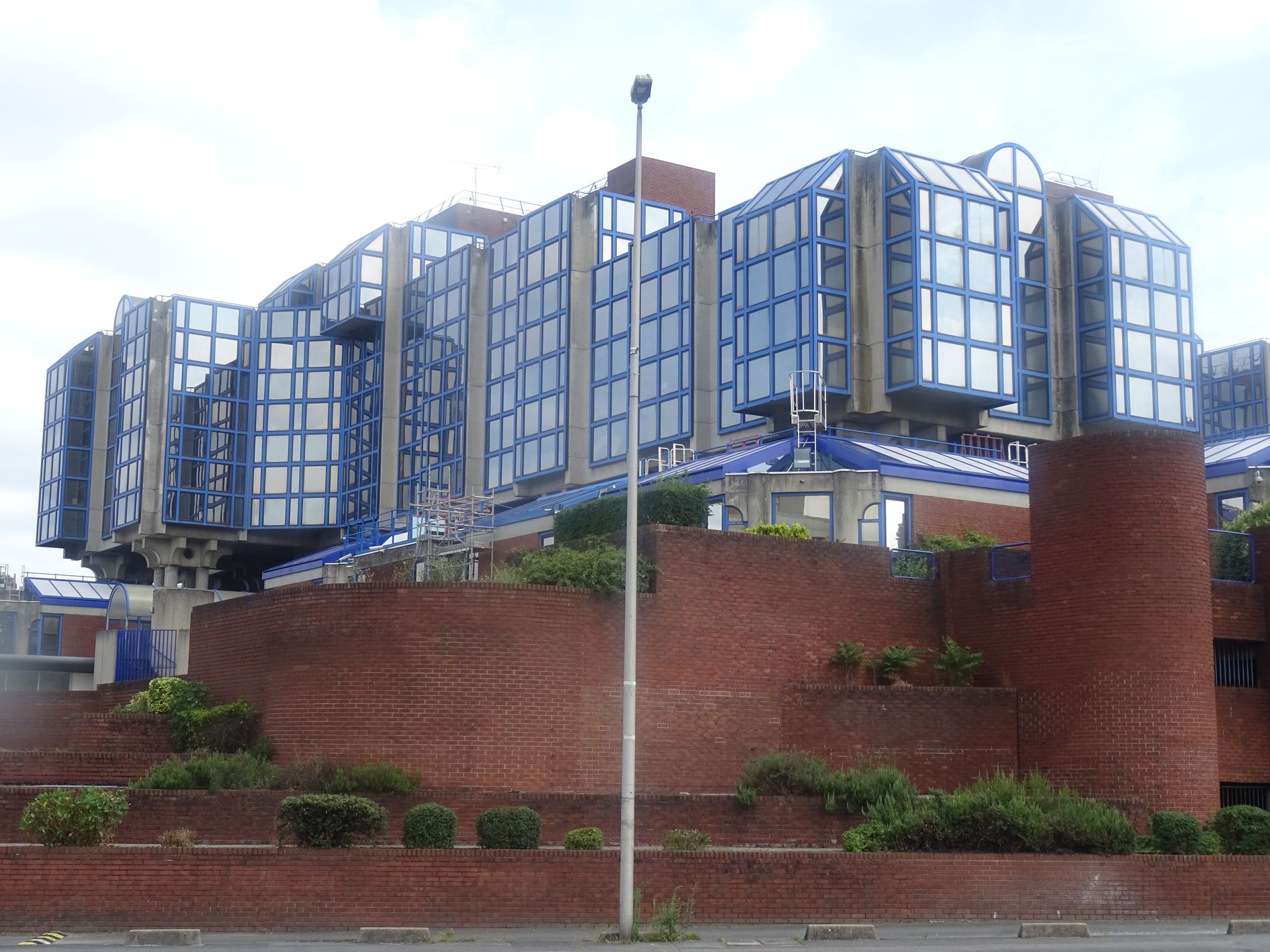 Bâtiment moderne avec des panneaux de verre à cadre bleu sur une base en briques rouges et des murs de soutènement étagés.