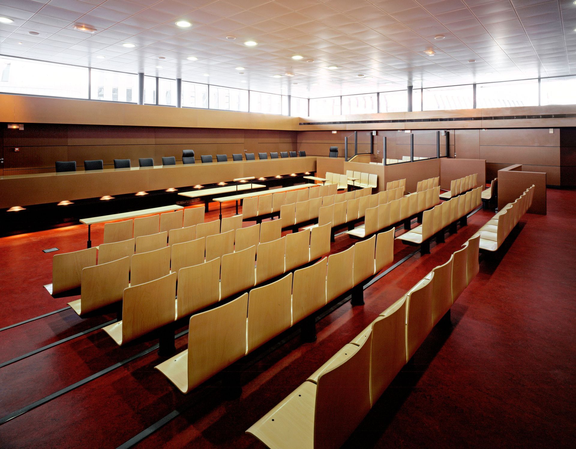 Auditorium avec des rangées de sièges en bois, de longues tables et un sol rouge.