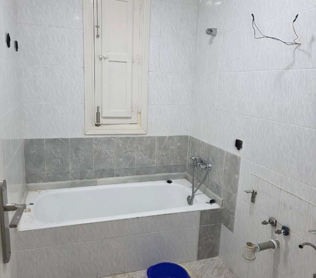 Baño pequeño sin terminar con bañera blanca, azulejos grises, tuberías a la vista y pintura de pared agrietada.