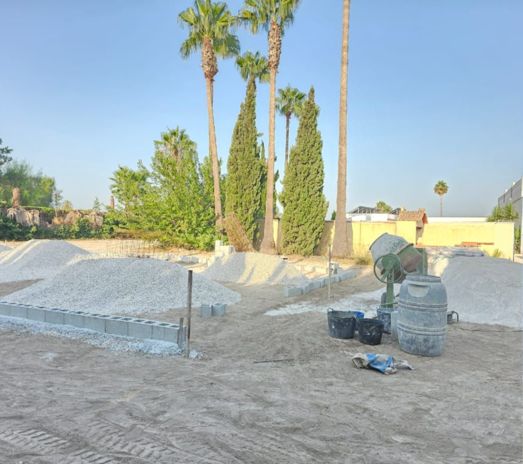 Obra en construcción con palmeras, montones de grava y una hormigonera en una zona abierta y arenosa.