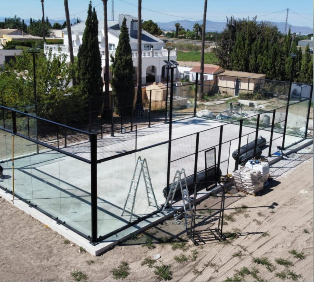 Pista deportiva exterior vallada en construcción, con base de hormigón blanco y estructura metálica negra