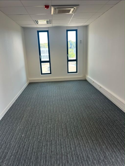 Bureau vide avec une moquette grise, des murs blancs et deux fenêtres étroites au fond.