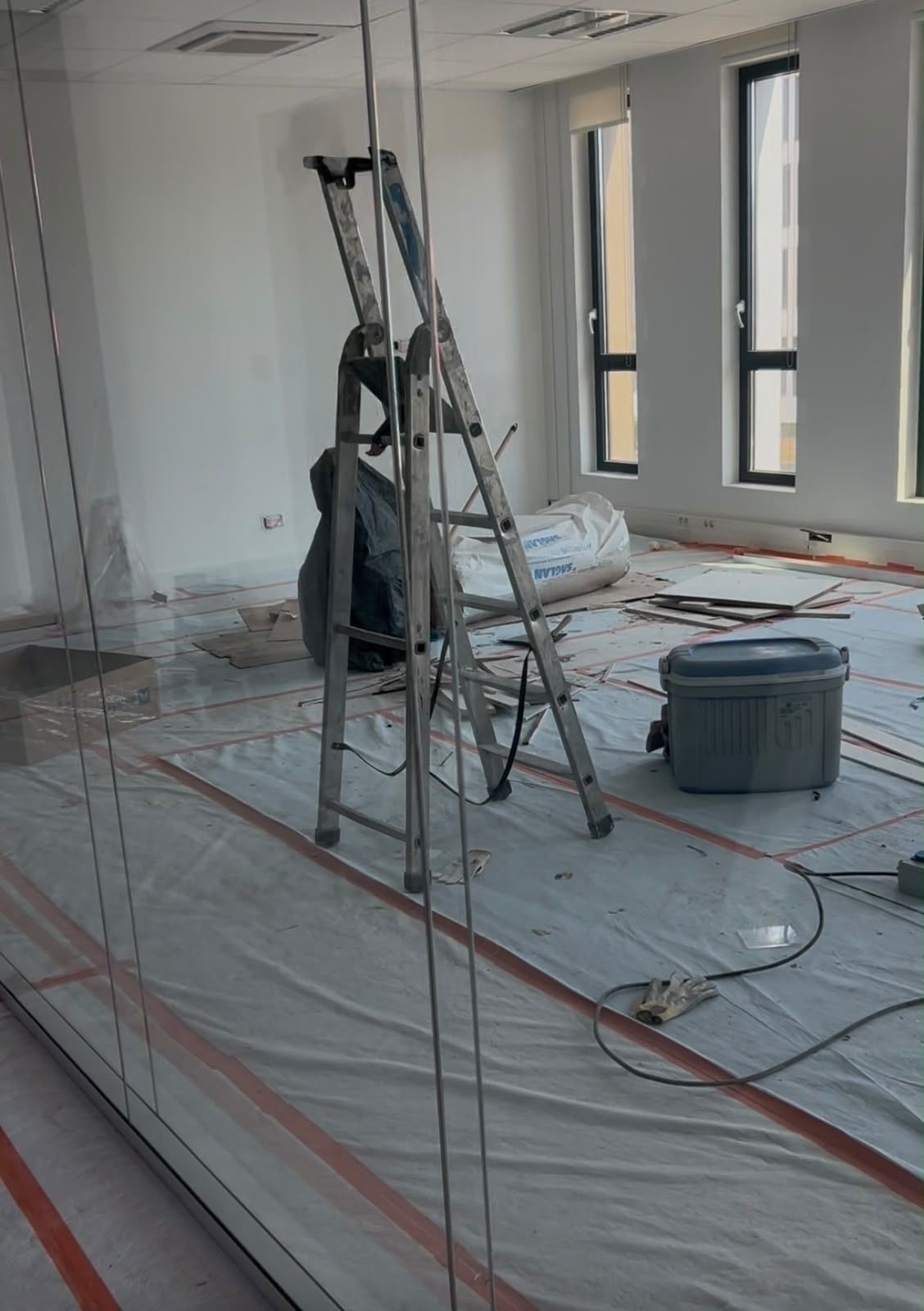 Pièce en rénovation avec des échelles, un sol protégé, des rallonges orange et de grandes fenêtres