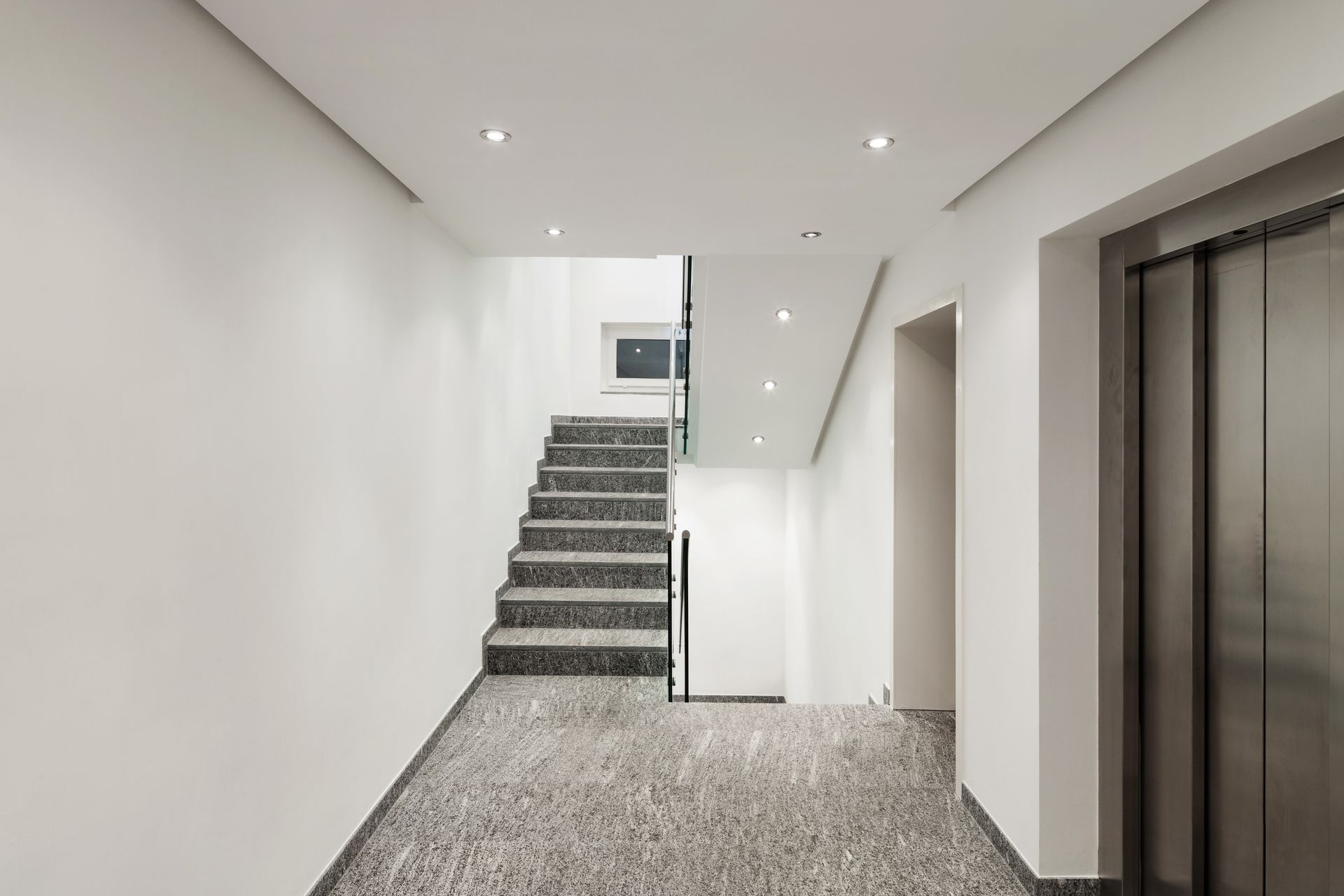 Couloir blanc minimaliste avec un escalier et un ascenseur, éclairé par des spots encastrés au plafond.