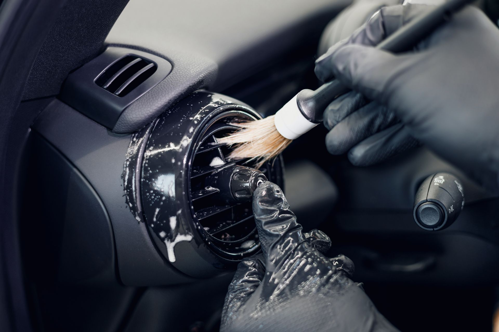Des mains gantées nettoient une bouche d’aération du tableau de bord d’une voiture avec une brosse et un chiffon en microfibre