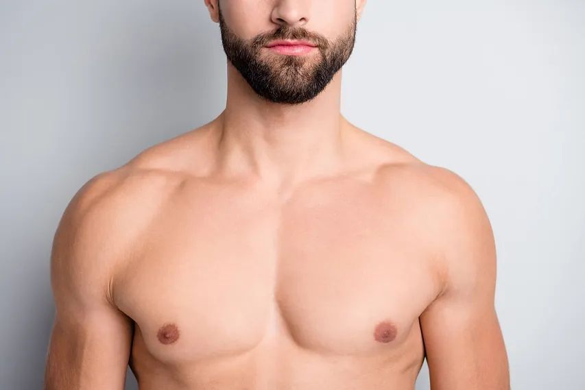 Hombre con barba y pecho desnudo, mostrando músculos pectorales definidos, sobre un fondo gris simple.