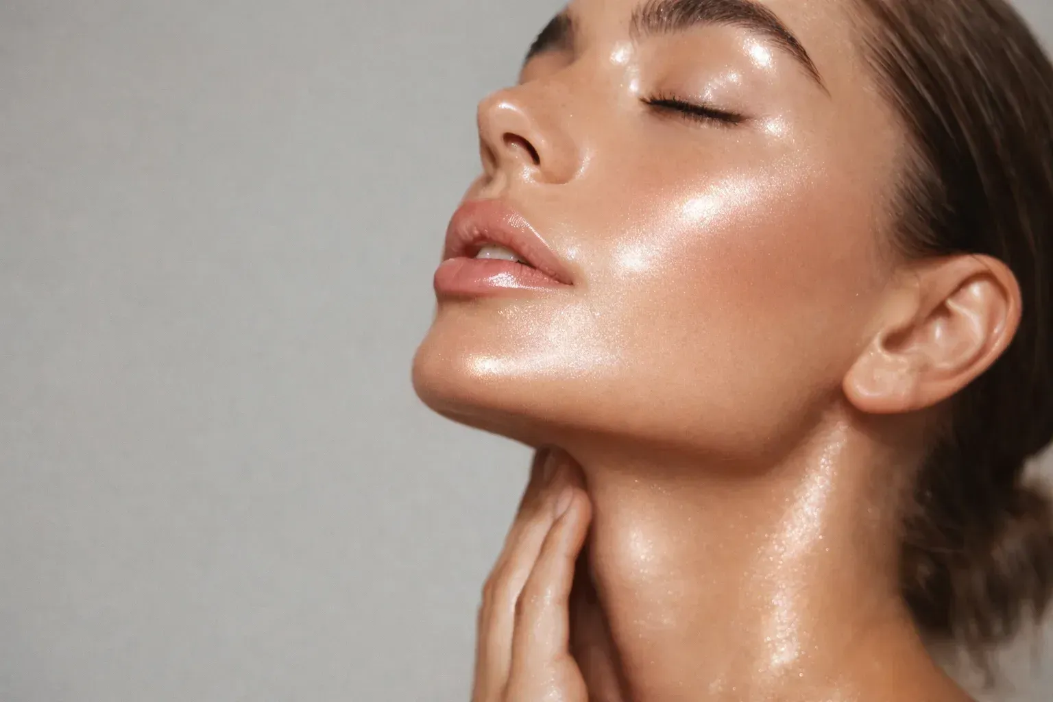 Mujer con piel brillante, ojos cerrados, mano en el cuello, sobre un fondo gris.
