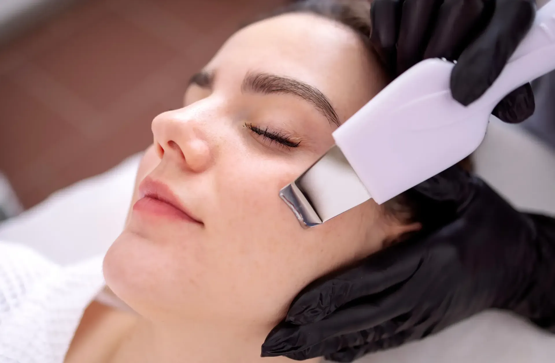Mujer recibiendo tratamiento láser facial en una clínica, acostada, con gafas y guantes rosas.