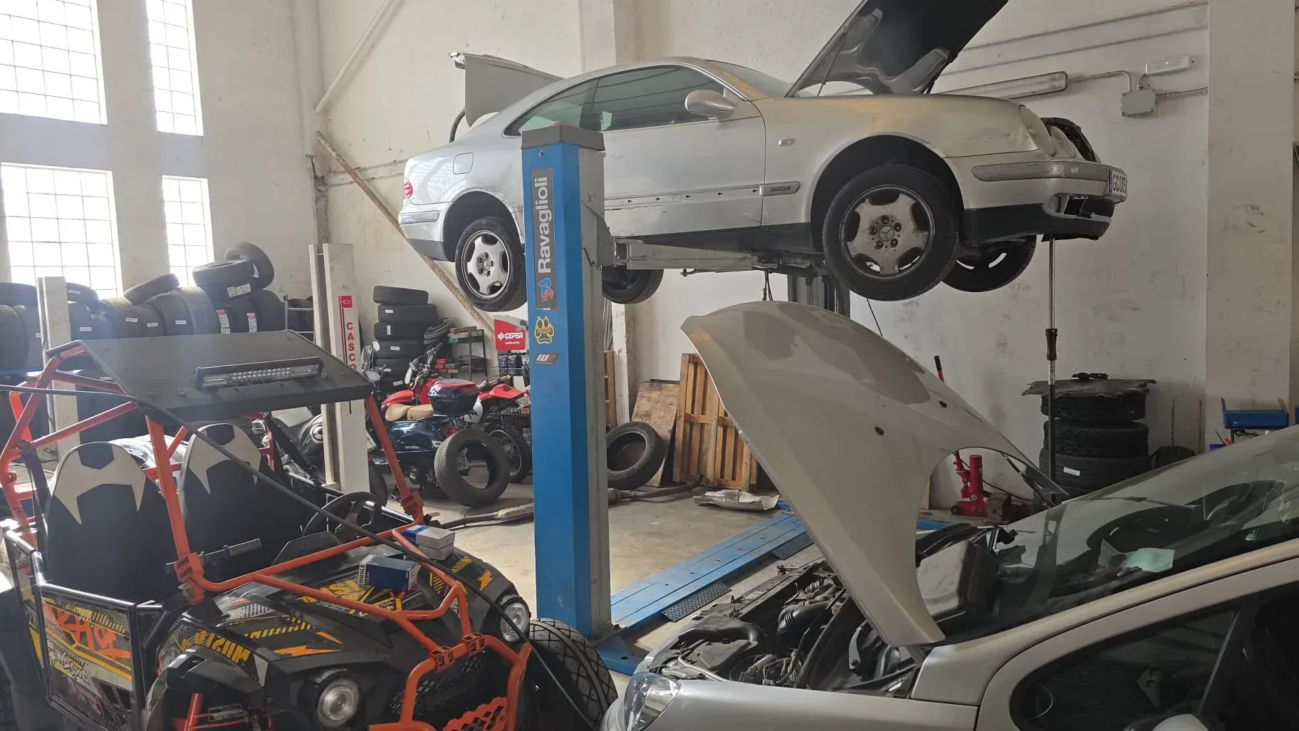 Un automóvil es levantado en un taller de reparación de automóviles junto a otro automóvil y un buggy en primer plano.
