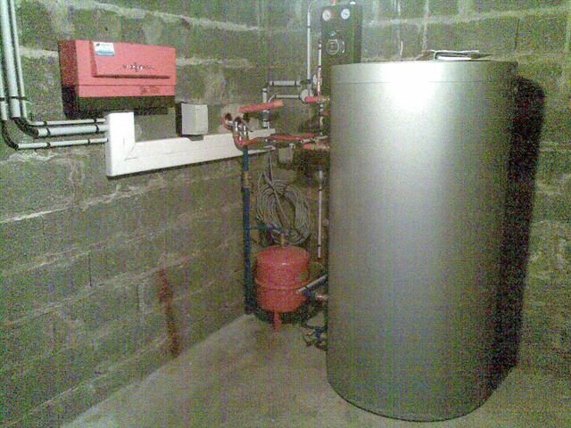 Une chaufferie avec un grand réservoir argenté, un vase d'expansion rouge et un panneau de commande