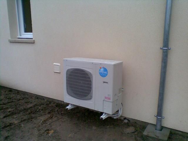 Unité de climatisation extérieure fixée sur un mur beige clair près d'une fenêtre et d'une descente de gouttière