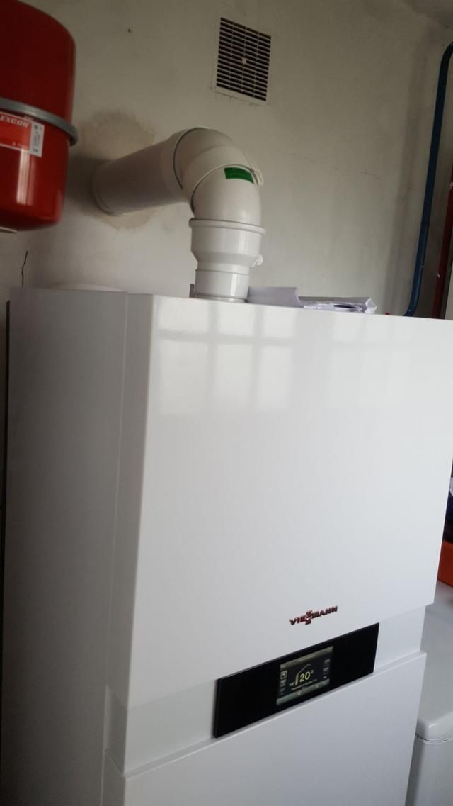 Chaudière blanche avec tuyau d'évacuation, fixée sur un mur blanc ; vase d'expansion rouge