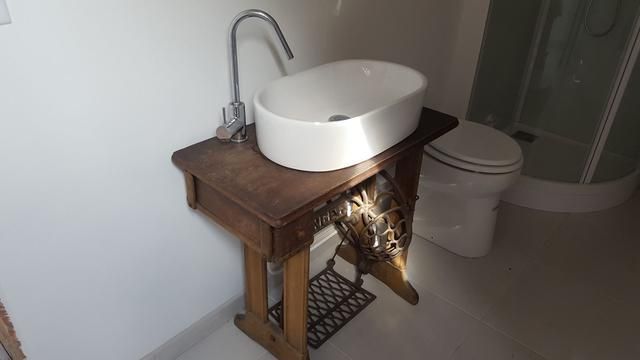 Lavabo avec vasque ovale blanche posée sur une ancienne table de machine à coudre, toilettes et cabine de douche