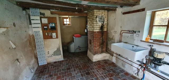 Intérieur d'une vieille pièce rustique avec un sol en tomettes, un évier, un tableau électrique mural et une chaudière