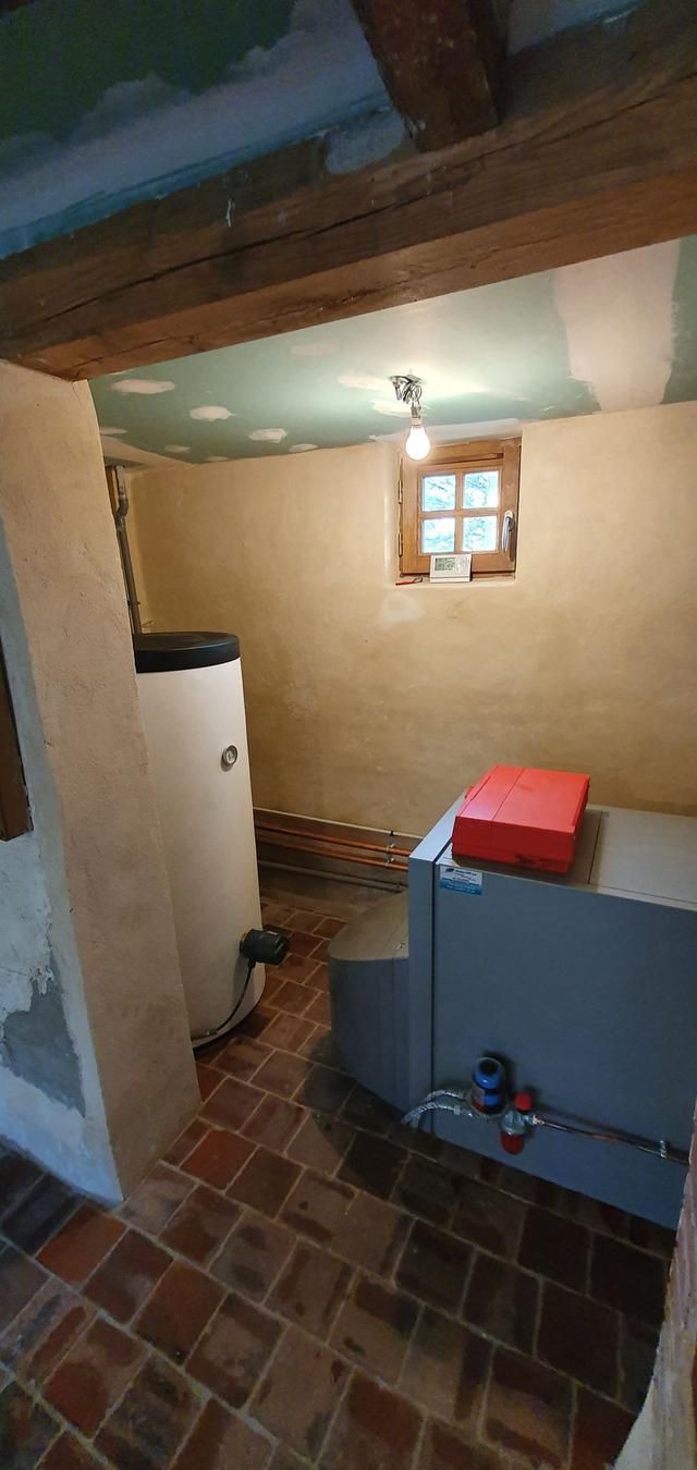 Un sous-sol avec chaudière, tomettes au sol, murs beiges et poutres apparentes