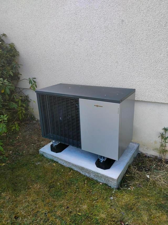 Unité de pompe à chaleur sur une base en béton, montée sur des isolateurs de vibrations noirs, à côté d'un mur