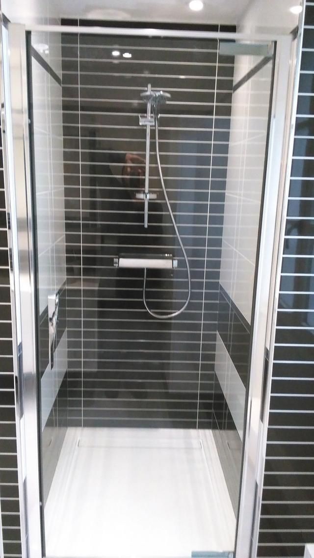 Cabine de douche avec porte en verre et cadre argenté, murs carrelés noirs et sol blanc