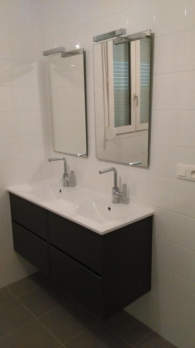 Salle de bains avec double vasque, deux miroirs avec éclairage intégré, meuble-lavabo foncé. Murs carrelés blancs, sol gris