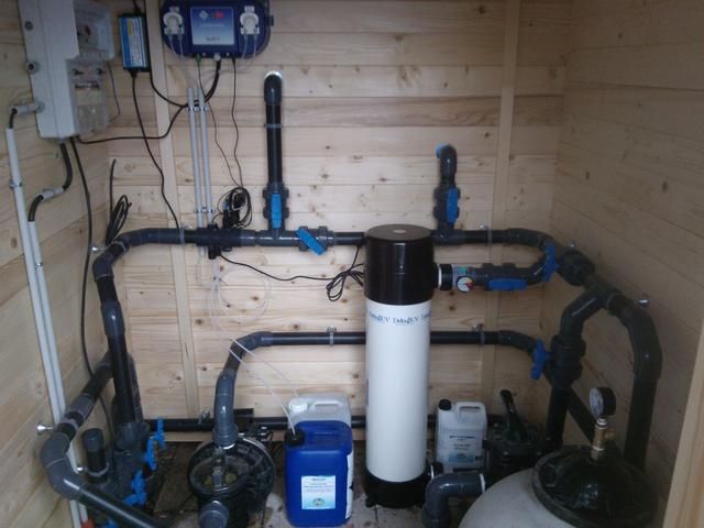 Local technique de piscine avec tuyauterie, filtre, pompes et panneau de commande, le tout adossé à des panneaux de bois
