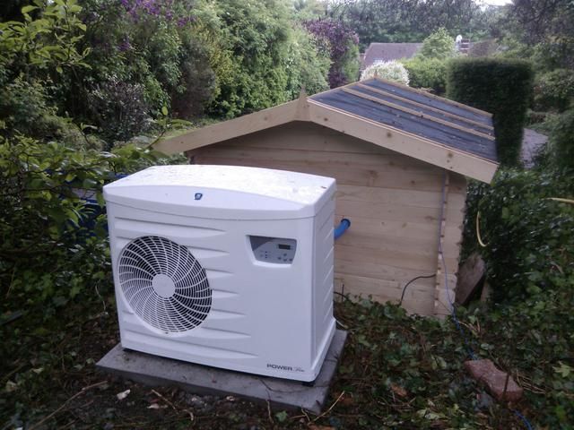 Unité de pompe à chaleur blanche sur une dalle de béton, à côté d'un abri de jardin en bois, dans un jardin