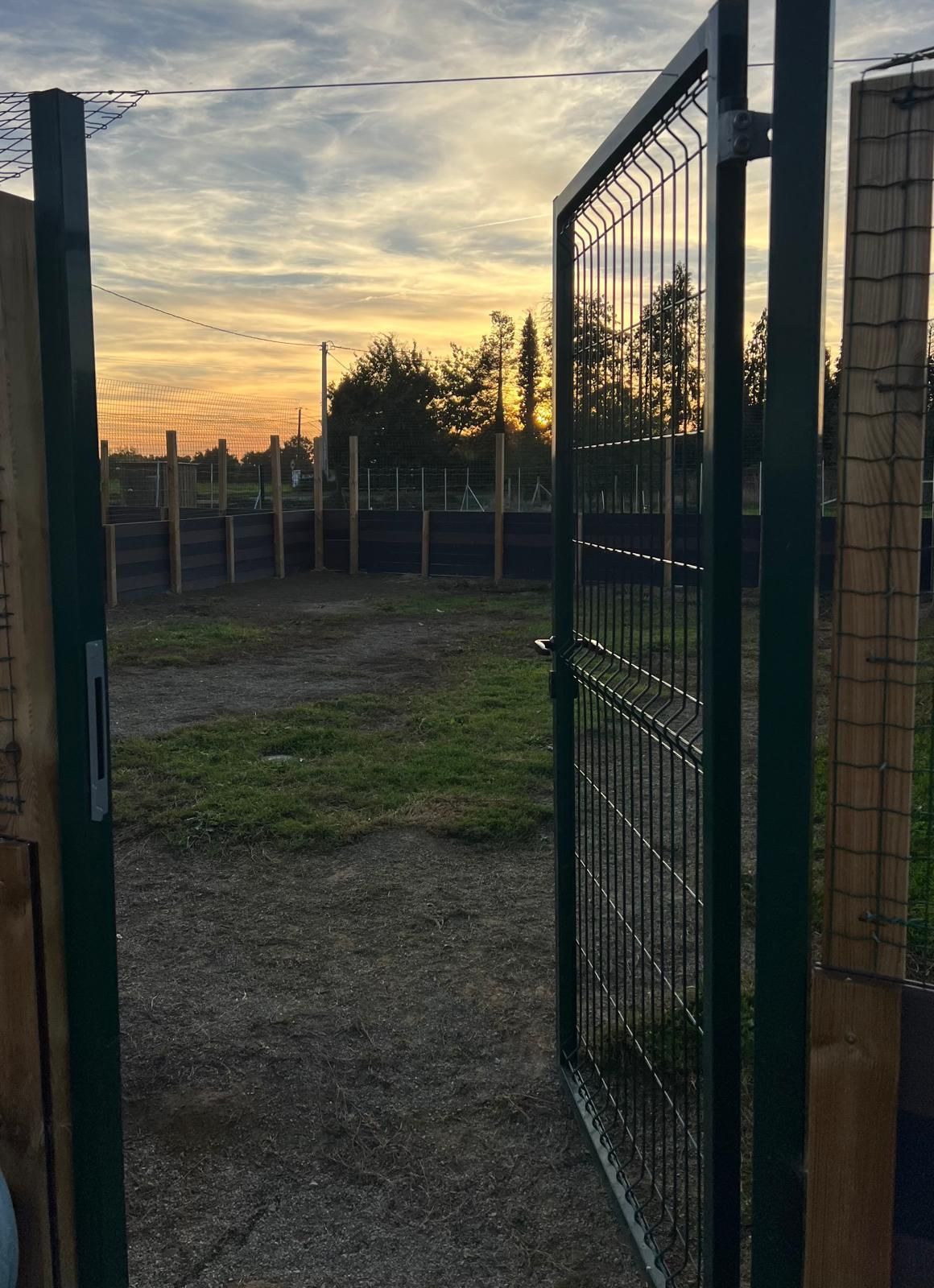Portail métallique ouvert donnant sur un espace herbeux avec clôture et un coucher de soleil.
