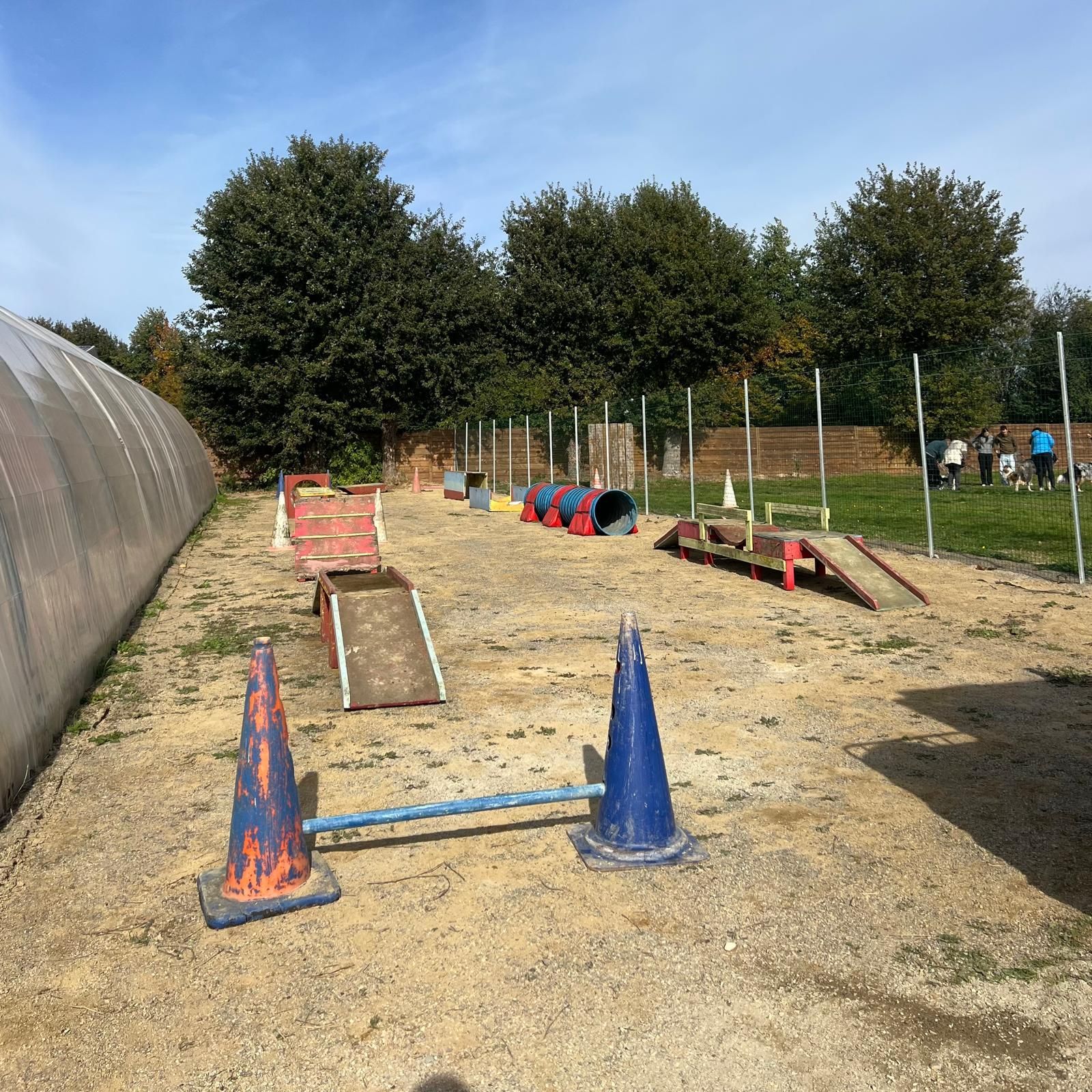 Parcours d'agilité canine : tunnels, rampes et sauts sur gravier. Des personnes en arrière-plan.