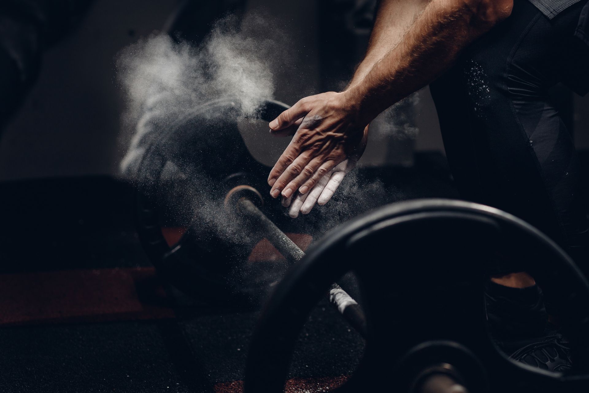 Des mains qui applaudissent, avec de la poussière de craie près d’une barre d’haltères dans une salle de sport sombre.