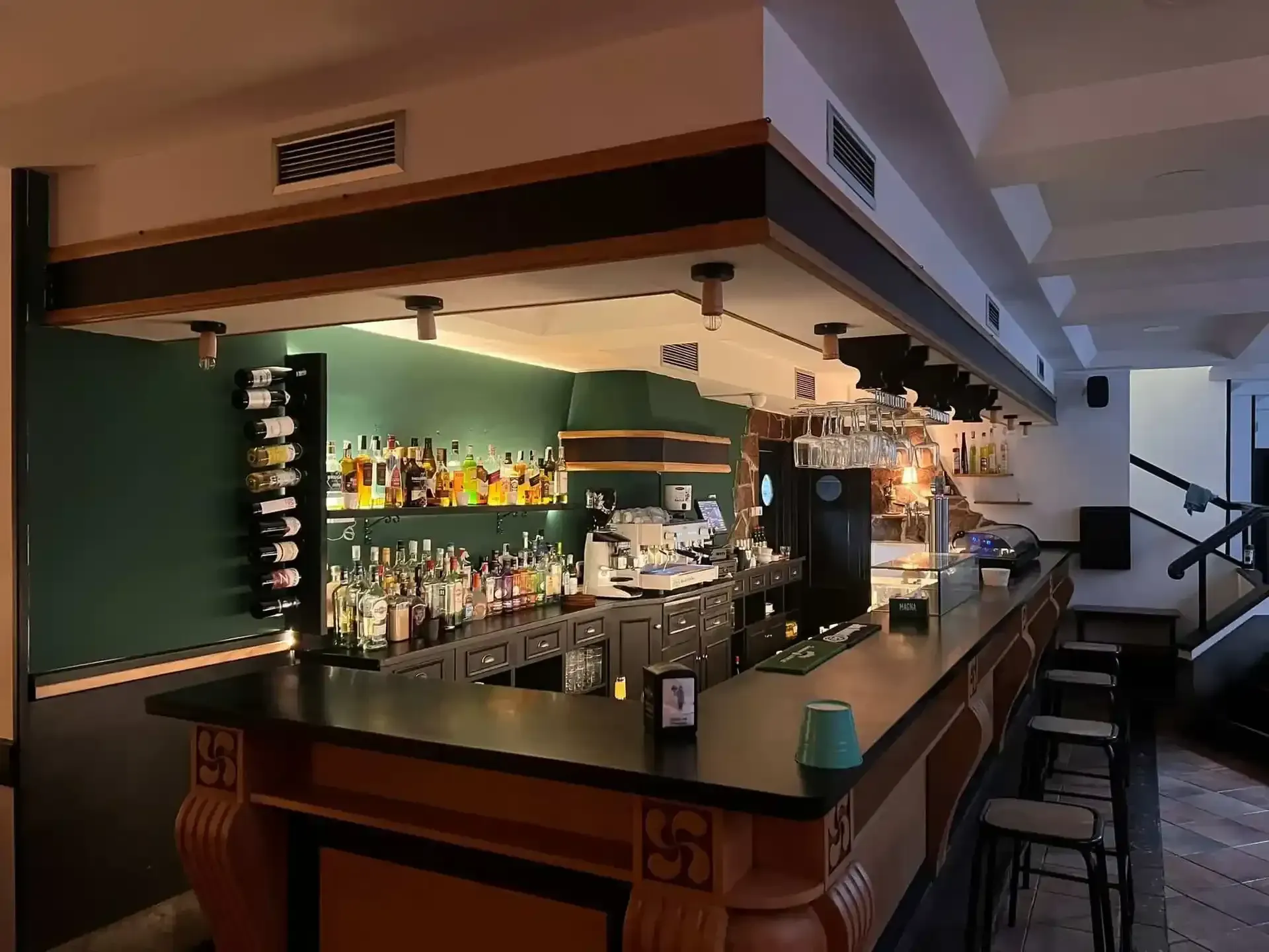 Interior de bar con botellas, cristalería y taburetes. Detalles en verde y madera, iluminación cenital.