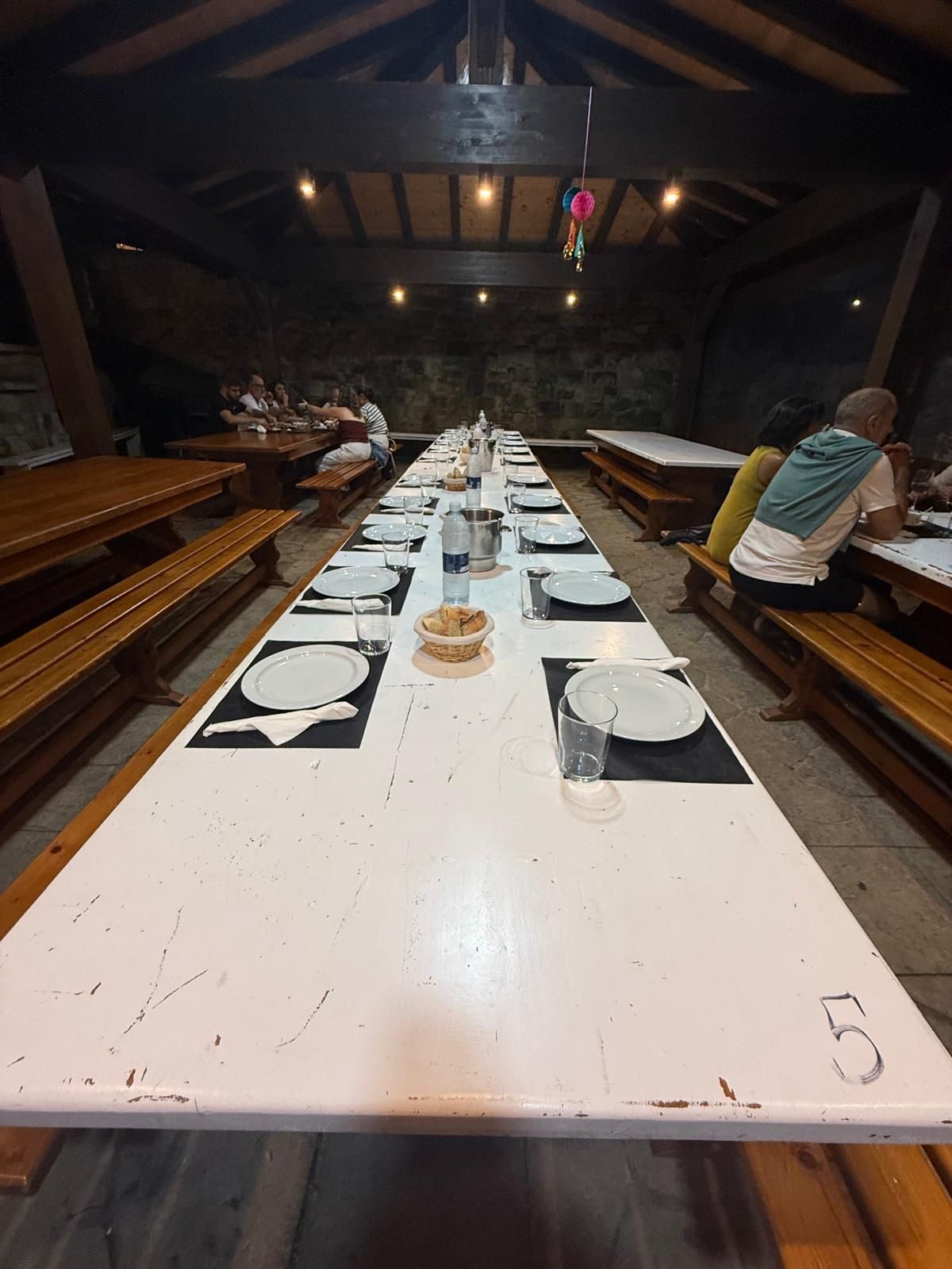 Mesa larga de madera dispuesta para comer en un espacio interior rústico con gente sentada en las mesas.