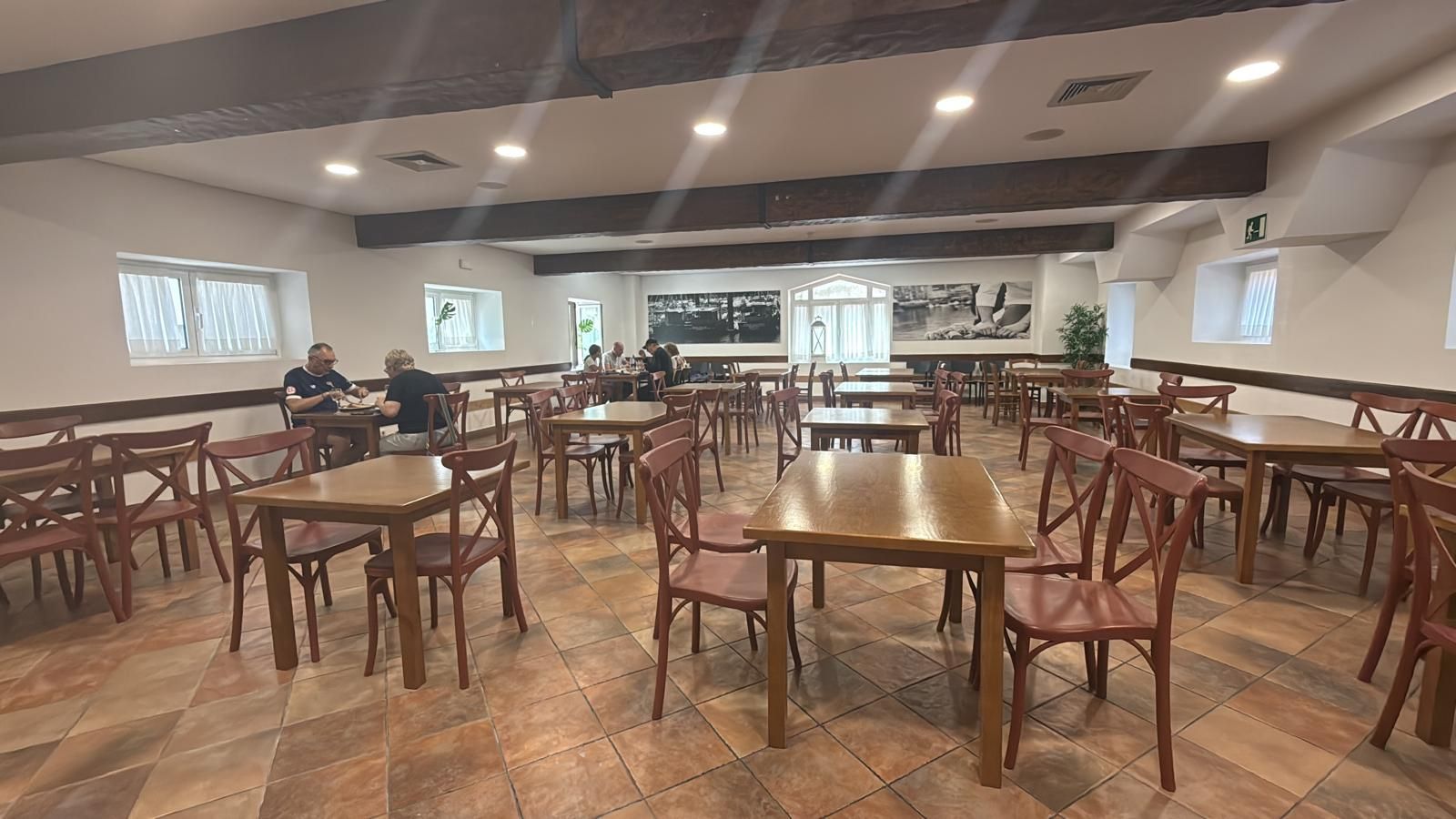 Comedor con mesas y sillas de madera. Personas sentadas en algunas mesas.