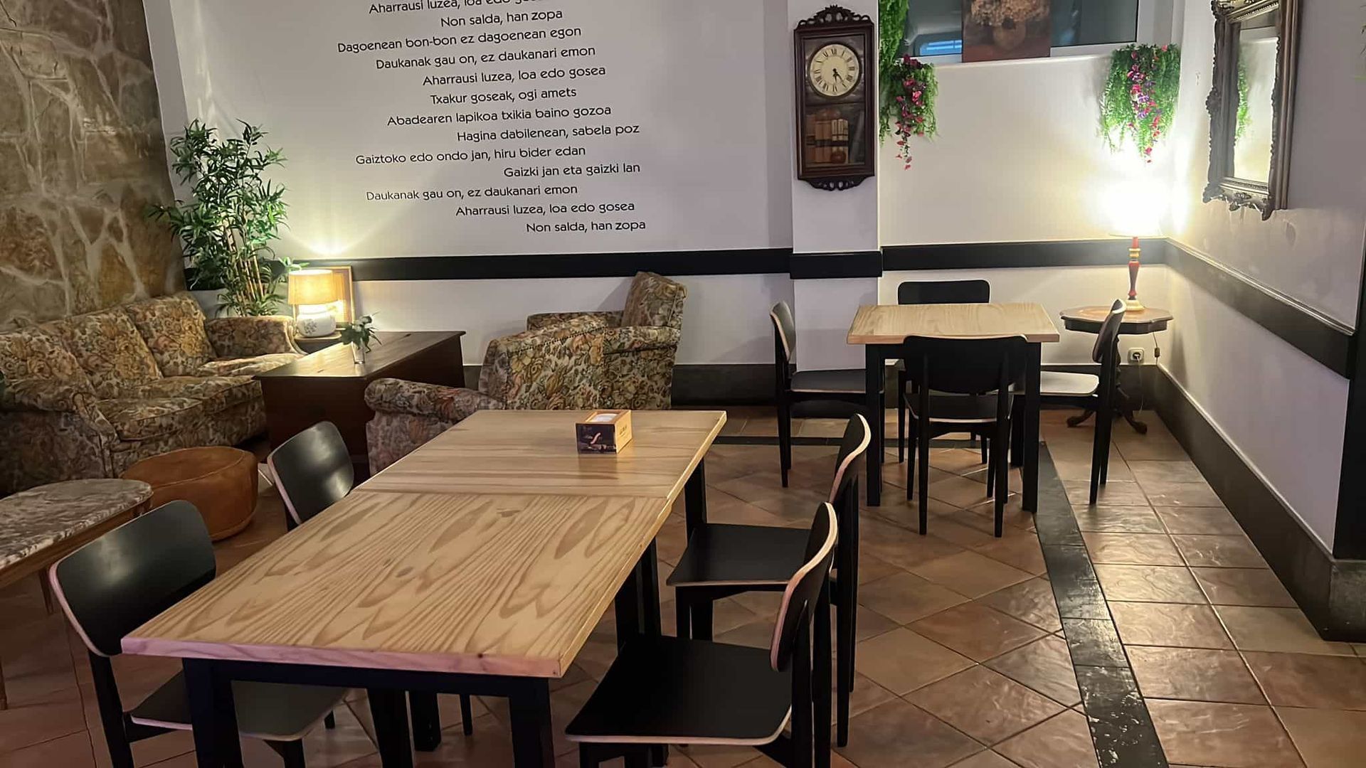 Interior de una cafetería con mesas, sillas, sofá, reloj y una pared decorativa.