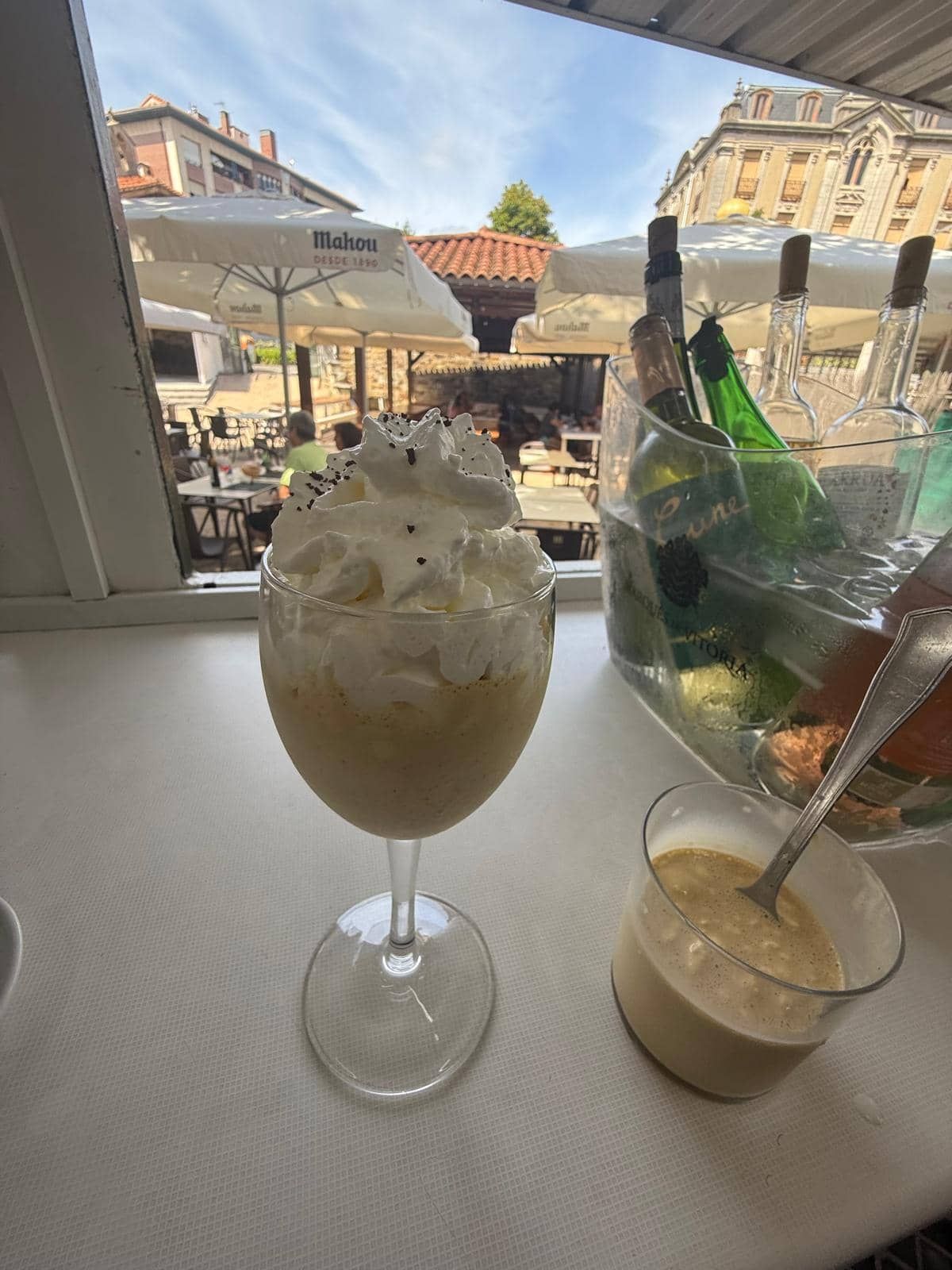 Un vaso de bebida cubierto con crema batida y una taza pequeña de acompañamiento sobre un mantel blanco; vista del patio exterior.
