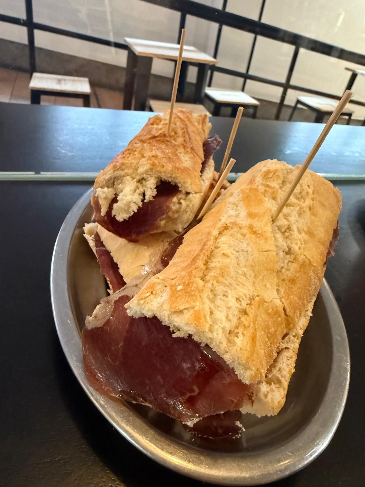 Dos sándwiches de jamón en una placa metálica, sujetos con palillos. Fondo borroso de mesas y sillas.
