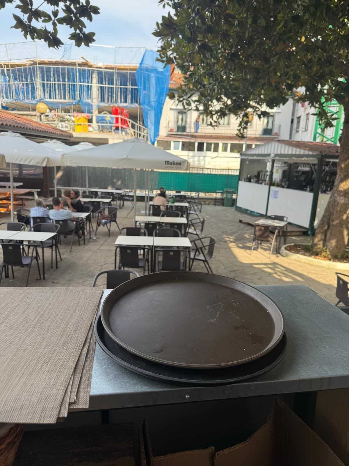 Café al aire libre con mesas vacías, grandes bandejas de metal en primer plano y un sitio en construcción al fondo.