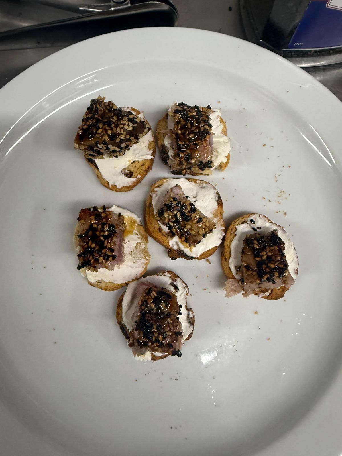 Seis pequeños aperitivos en un plato blanco: rodajas de pan tostado con queso crema y una cobertura oscura y desmenuzable.
