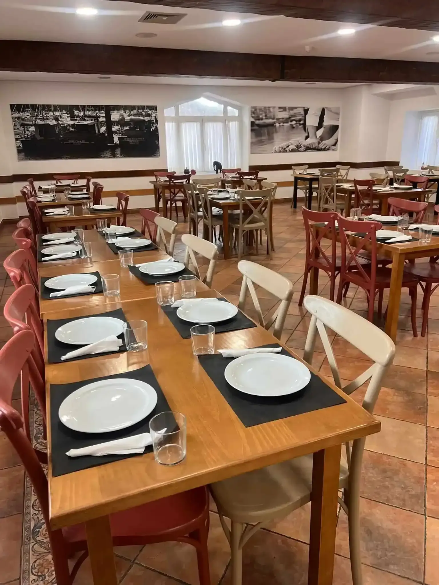 Interior de restaurante con mesas preparadas para comer; suelo de ladrillo, vigas de madera, sillas rojas y blancas, fotografías en blanco y negro.