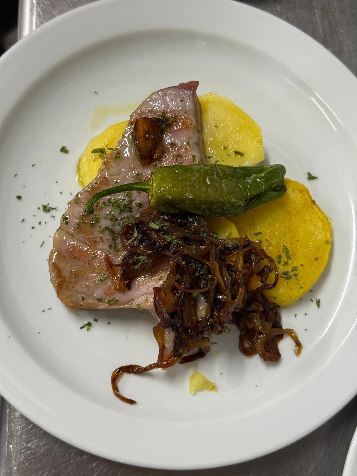 Bistec con patatas, cebollas caramelizadas y un pimiento verde en un plato blanco.
