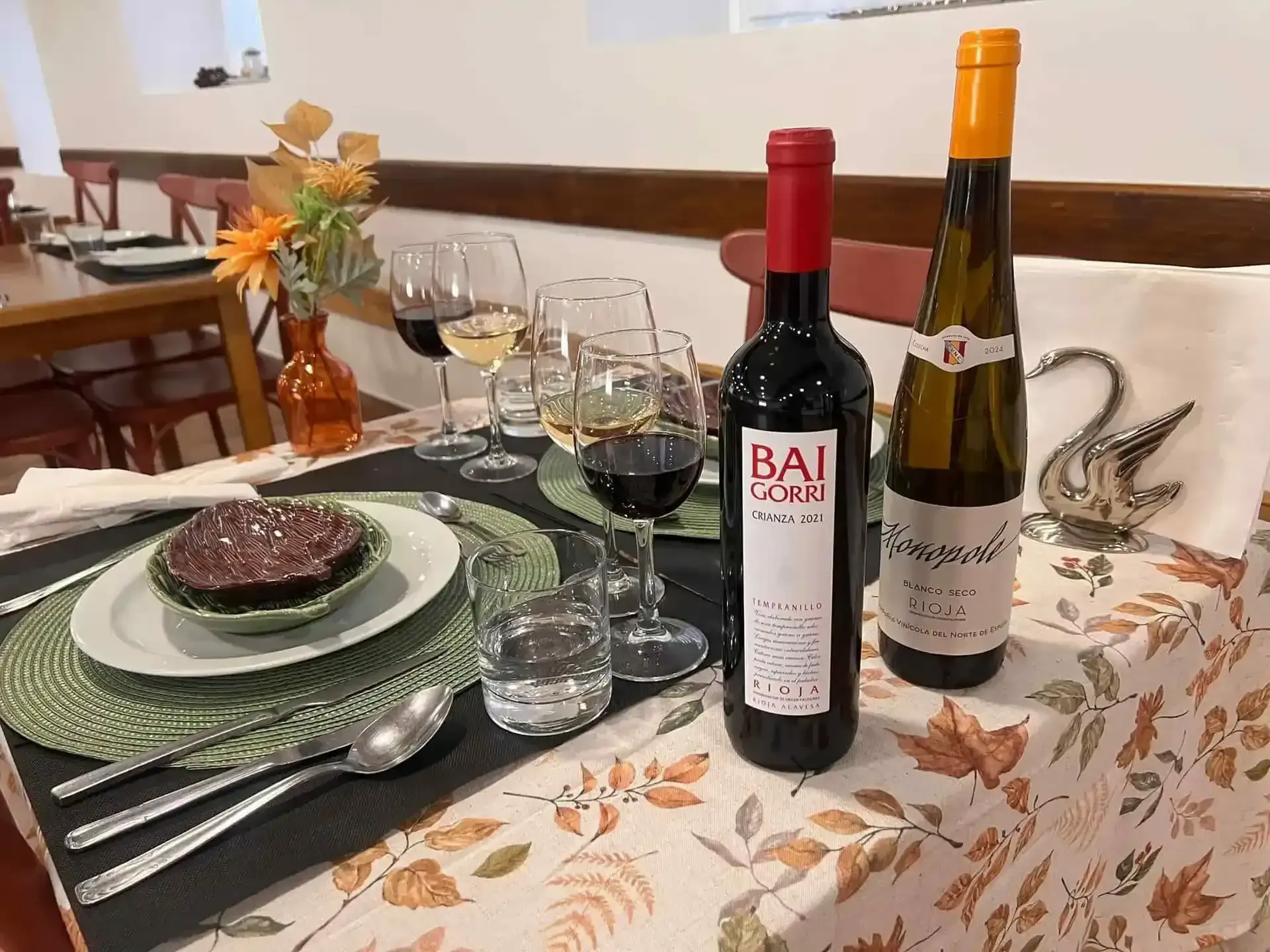 Mesa puesta para cenar con botellas de vino y copas, plato de comida y flores.