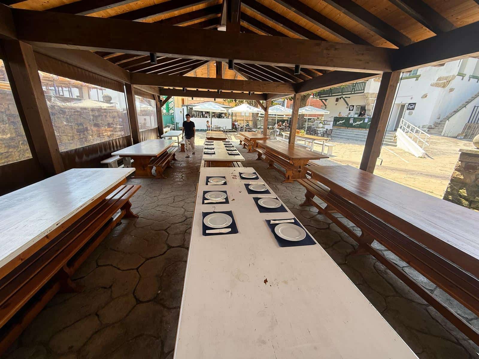 Comedor exterior con mesas largas dispuestas con platos, bajo una pérgola de madera.