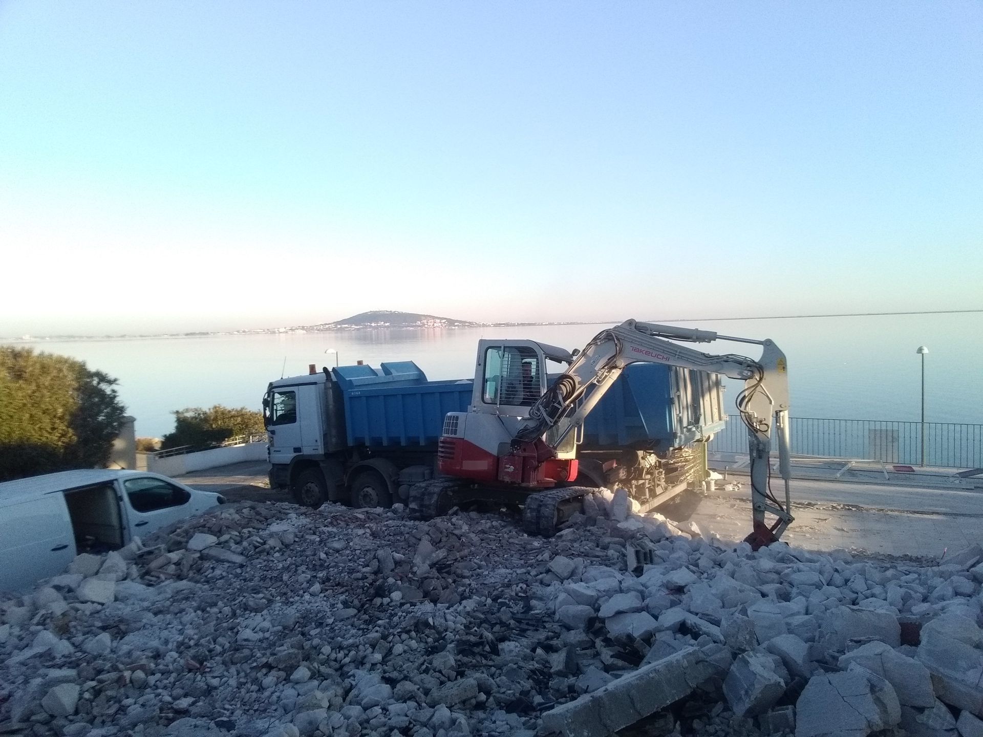 Une excavatrice charge des gravats dans un camion-benne près de l'eau. Ciel bleu.