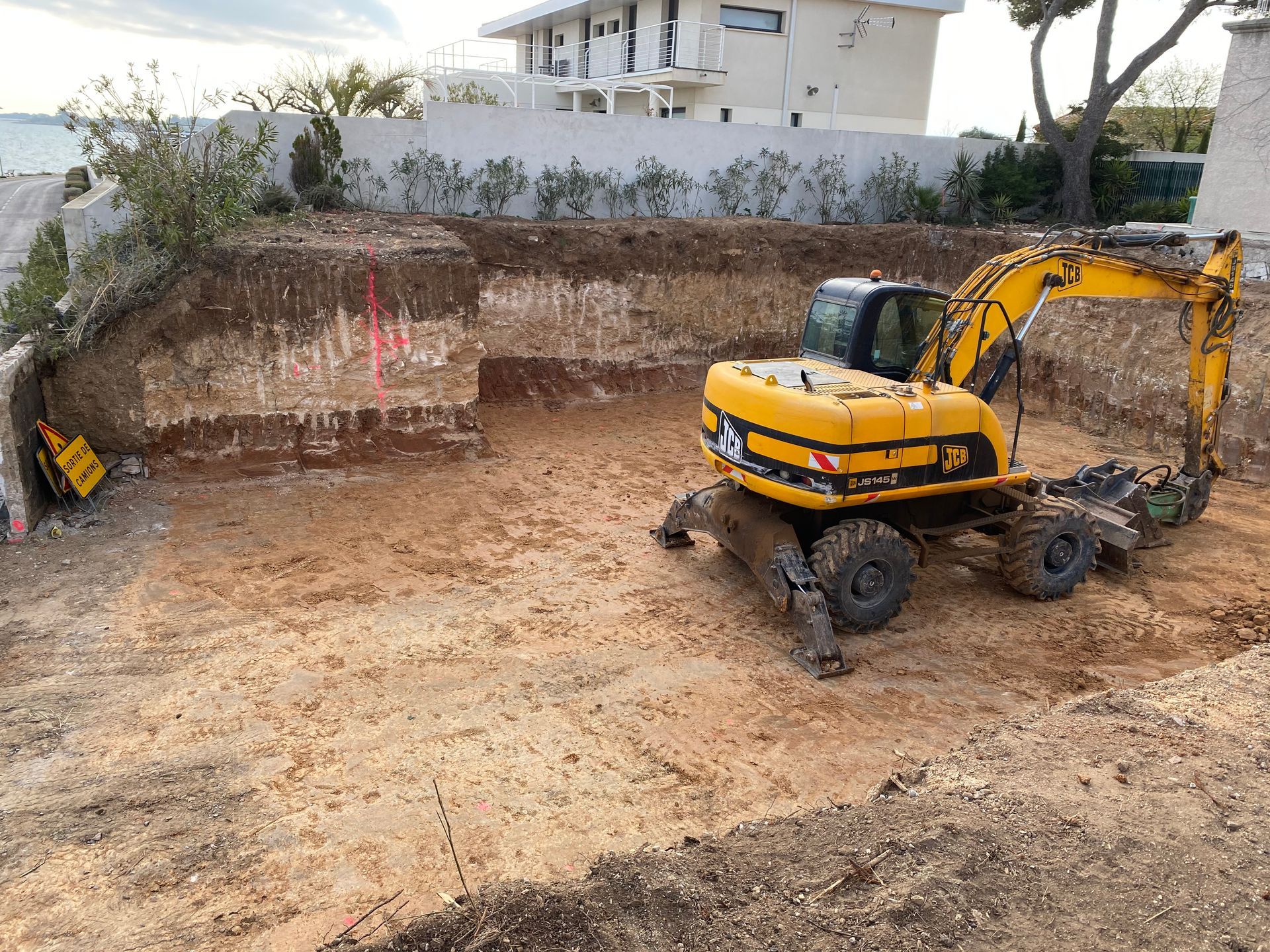 Une pelleteuse jaune creuse le sol sur un chantier. Un bâtiment, des arbres et une clôture sont visibles en arrière-plan.