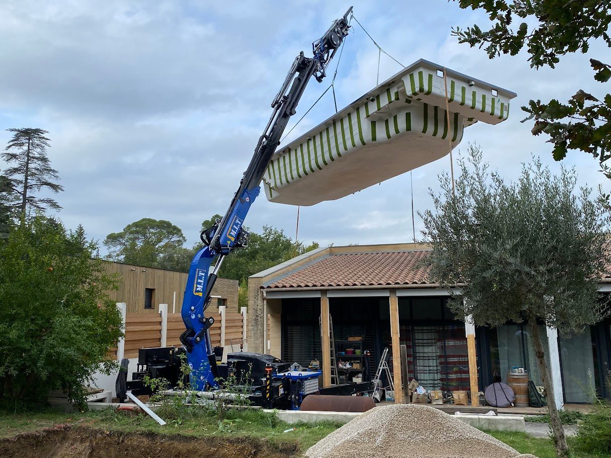 Une grue installe une piscine préfabriquée dans un jardin. Le cadre est ensoleillé, avec une maison et des arbres.