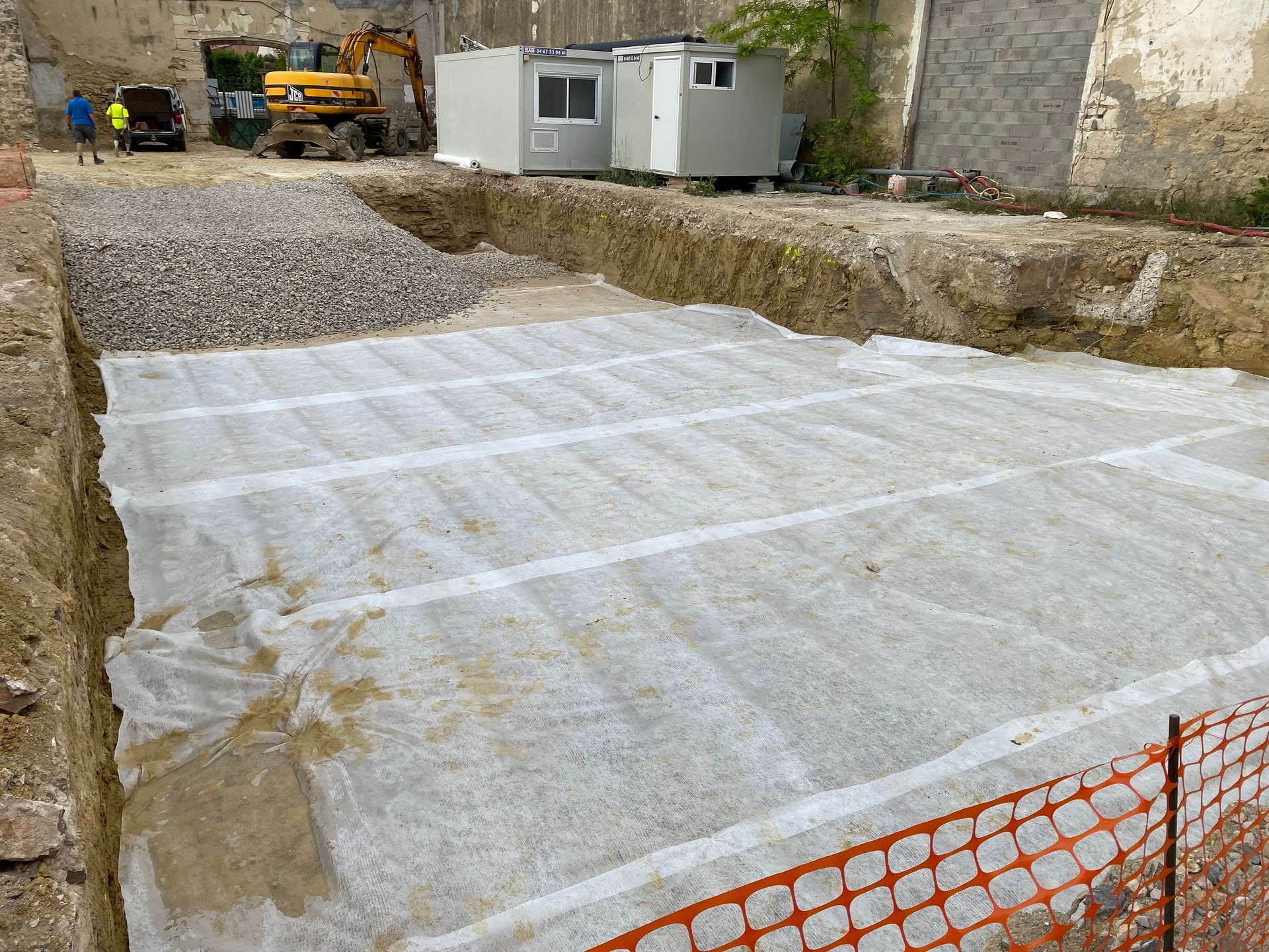 Chantier avec une excavation rectangulaire recouverte d'une bâche blanche. On aperçoit du gravier et des engins de chantier.