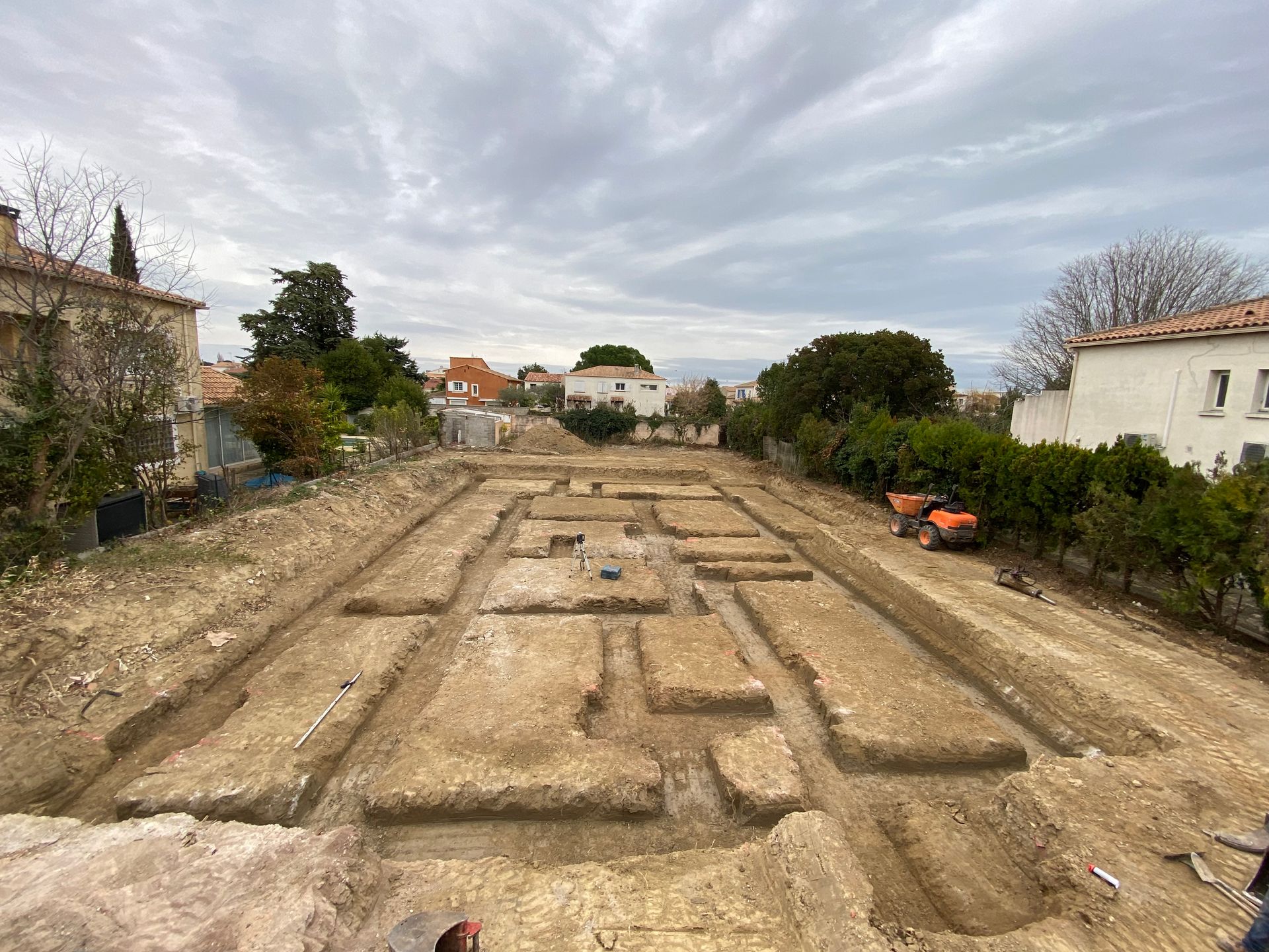 Site de fouilles archéologiques avec des tranchées rectangulaires révélant les fondations de bâtiments dans une zone résidentielle. Ciel couvert.