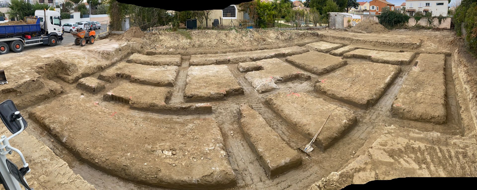 Fouilles archéologiques montrant les contours des fondations de bâtiments dans la terre, avec des engins de chantier en arrière-plan.
