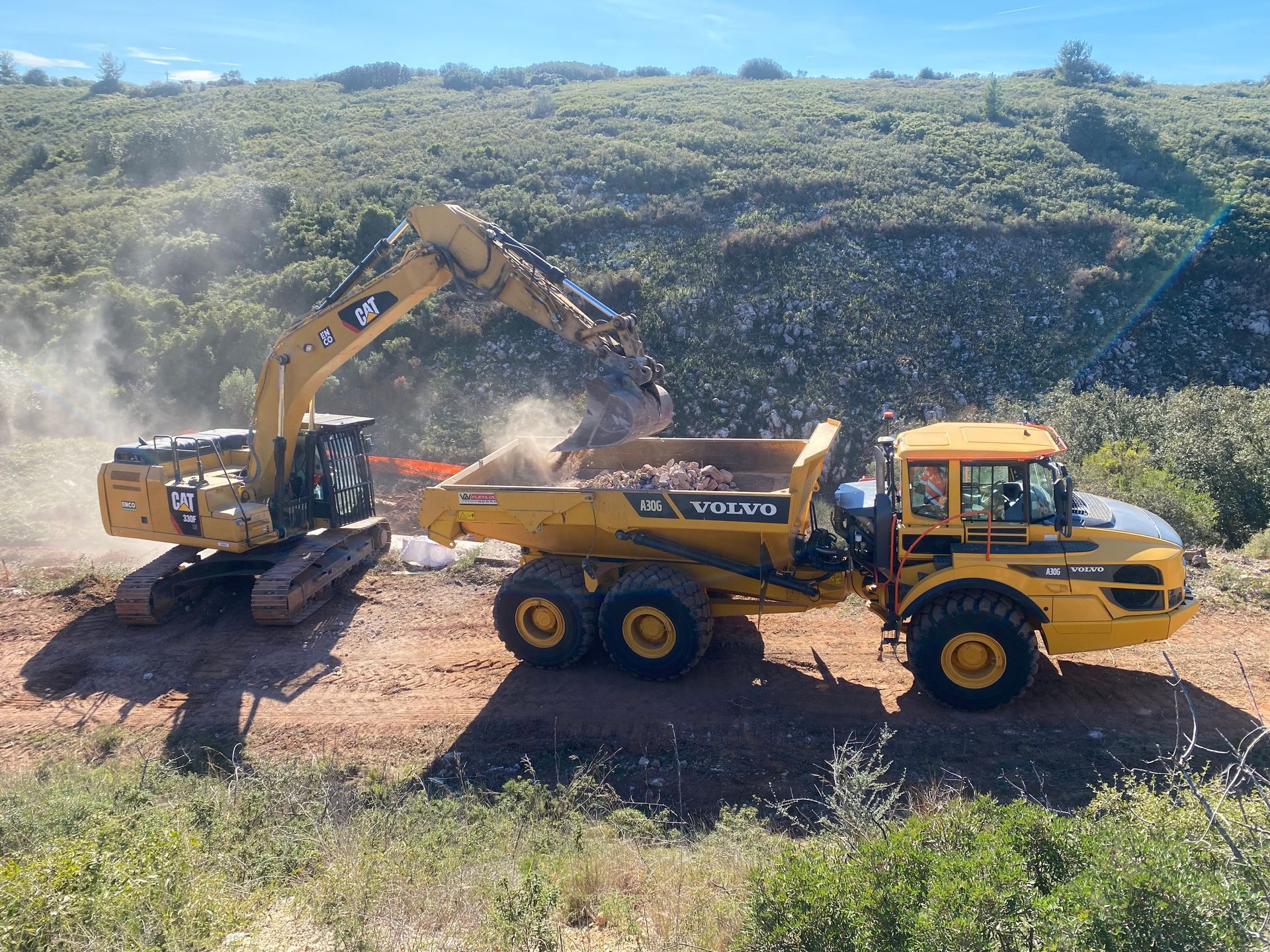 Une pelleteuse jaune charge des débris dans un camion-benne Volvo jaune sur un flanc de colline poussiéreux.