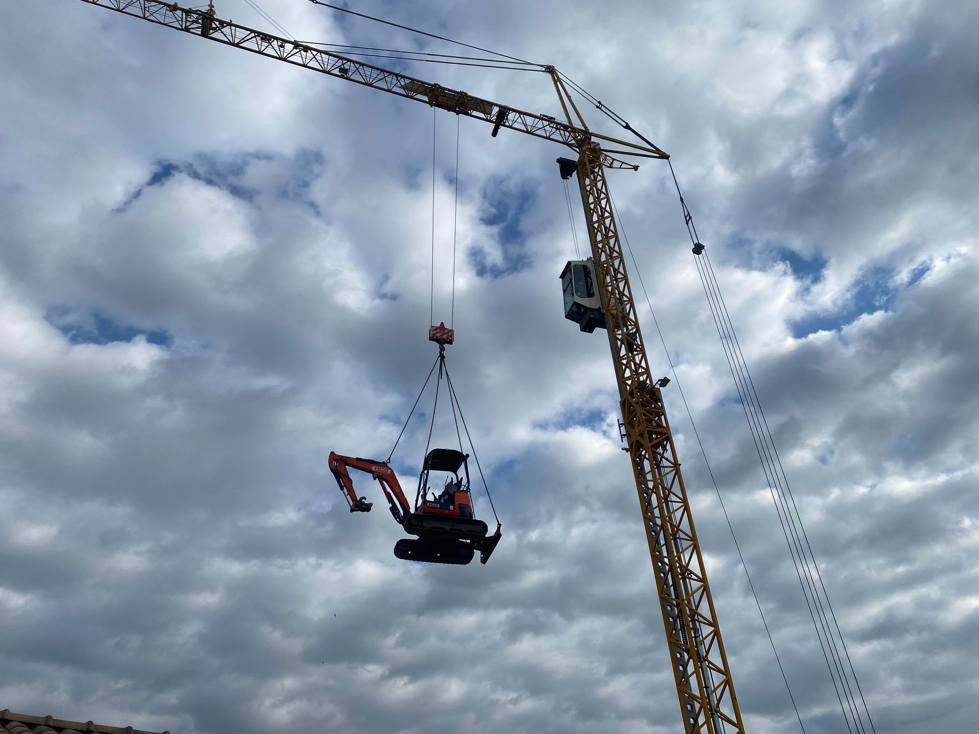 Une grue de chantier soulève une petite excavatrice rouge sur fond de ciel nuageux.